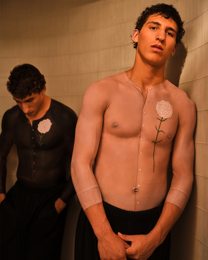 Twin brothers leaning against a grey wall, each with body paint on their chests which makes it appear that they are wearing button up sweaters
